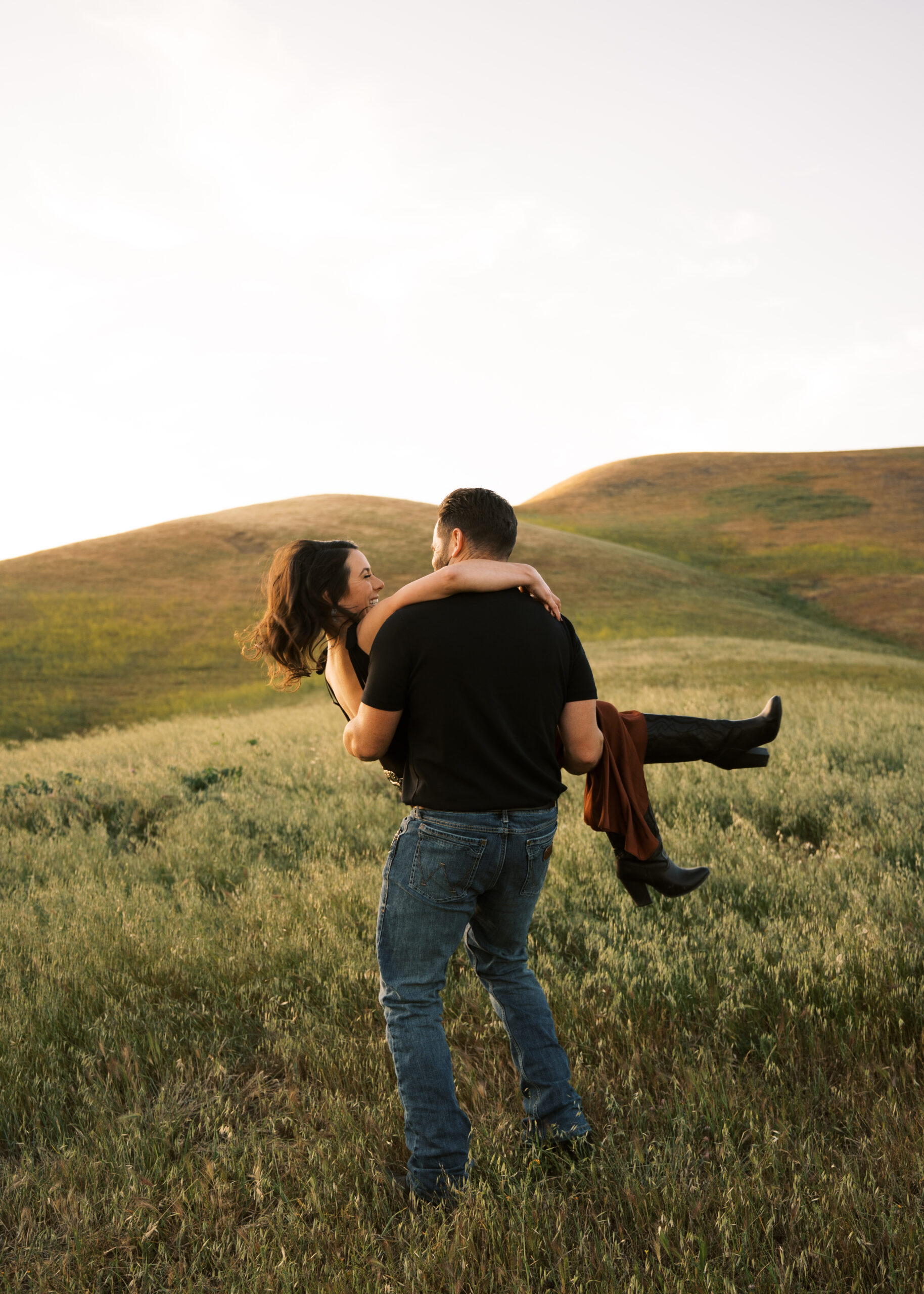 Man picking up woman and spinning her around in a field while she wears cowboy boots and a skirt at sunset