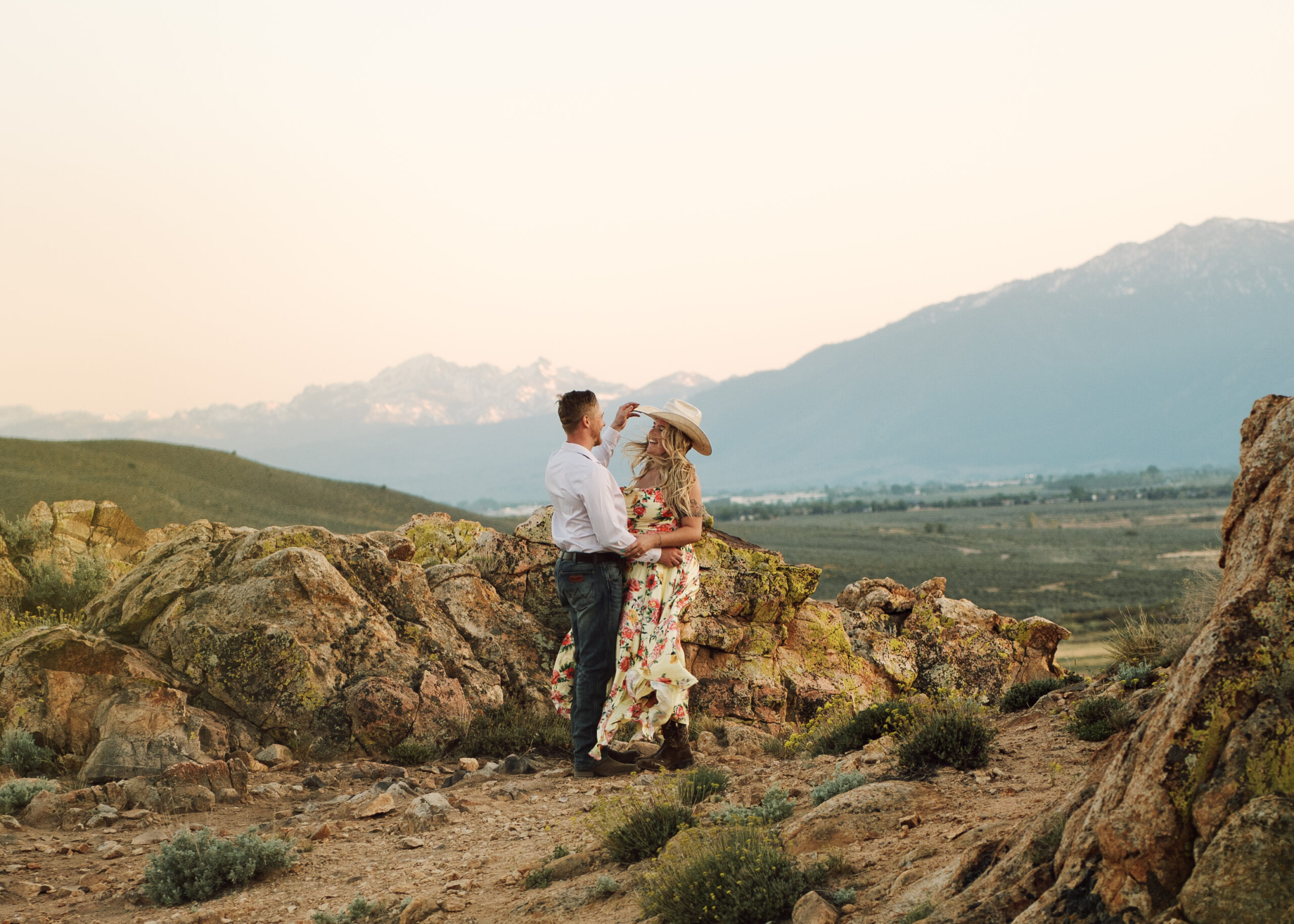 Fiancé offering his cowboy hat to his partner with a scenic mountain range in the background, a candid moment from windy engagement photos in Carson City, Nevada.