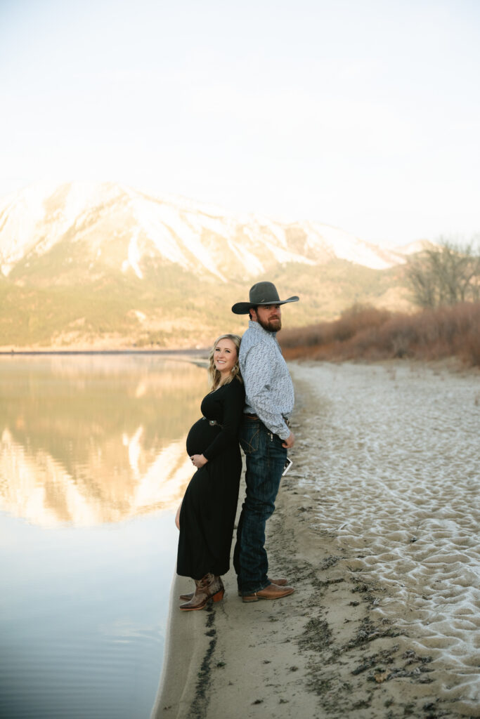 Man and pregnant woman standing back to back next to mountains and a lake