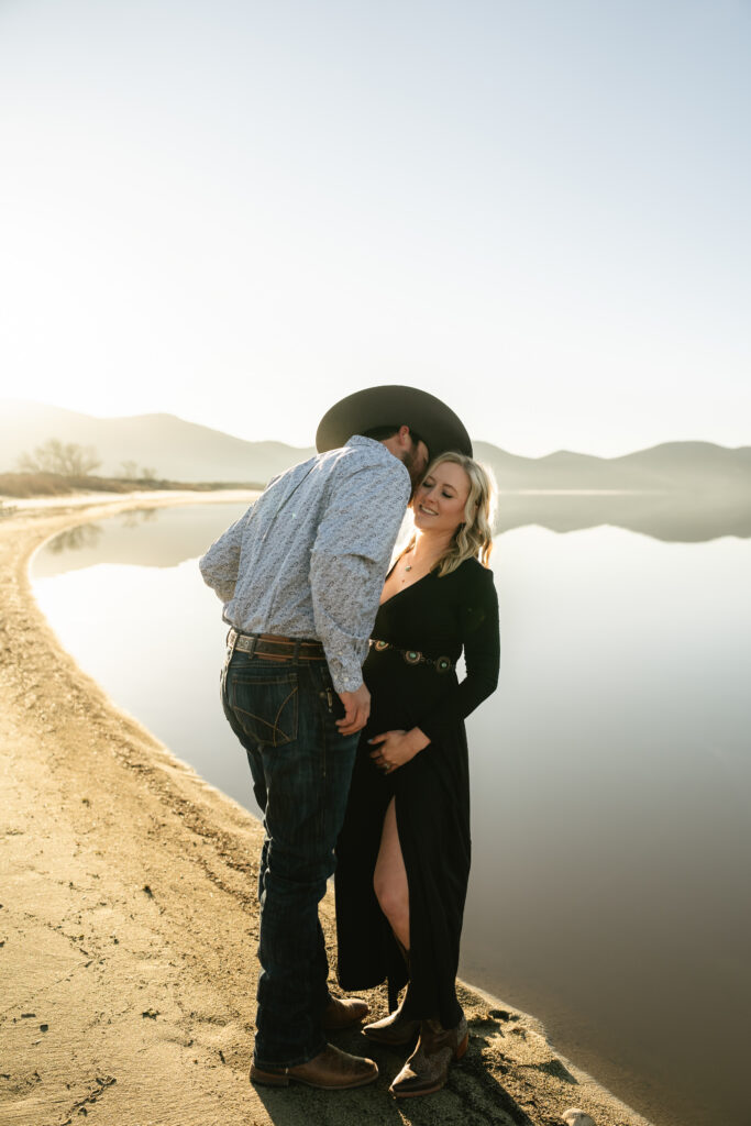Cowboy kissing pregnant wifes forhead by a lake and mountain background